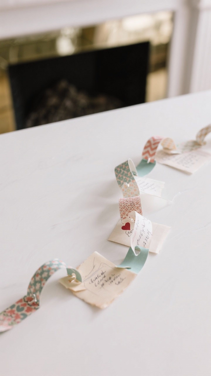 Overhead detail shot of love letter paper chains: alternating 1-inch strips of patterned scrapbooking paper and solids, with occasional vellum strips for lightness; each link features tiny handwritten notes and memories; linked loops draped across a white tabletop with a hint of a mirror edge and mantle in background; close focus on paper patterns, ink strokes, and the subtle translucence of vellum